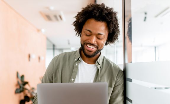 Young man in an office space smiling and looking down at laptop screen