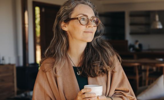 A woman wearing glasses indoors in a home setting holding a mug