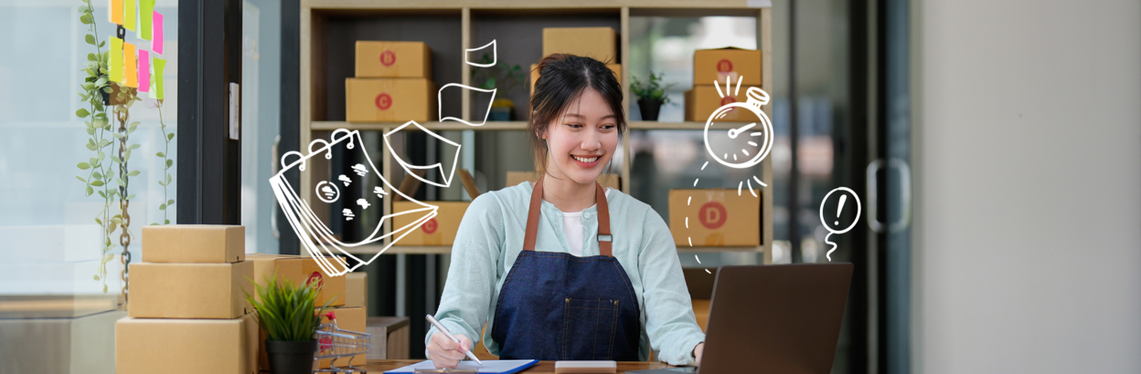 Female business owner using laptop at desk in her storefront with packages on a shelf behind her