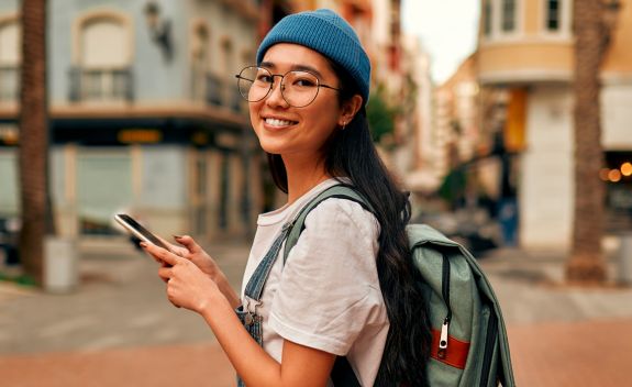 A smiling female student wearing a backpack and walking in city space while using mobile phone