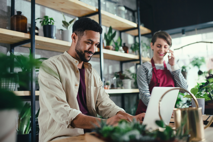 Man working on a laptop in a store selling potted plants, with shop assistant in the background on phone